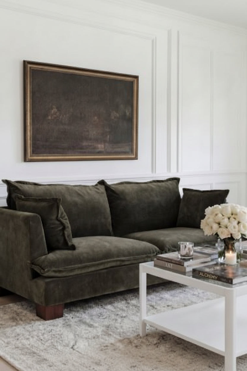Olive green sofa in a living room with a white wall, framed artwork, and a coffee table with decor items.